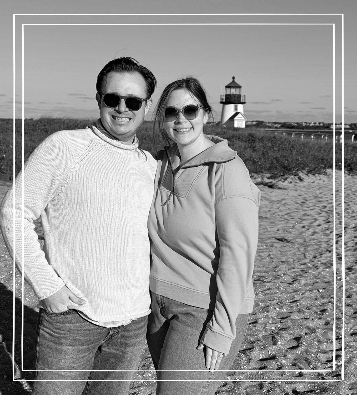 A black and white lifestyle photograph of a couple standing on a sandy beach with a lighthouse in the background. The man on the left wears a light-colored knit sweater and sunglasses, and the woman on the right wears a light-colored pullover hoodie and sunglasses.