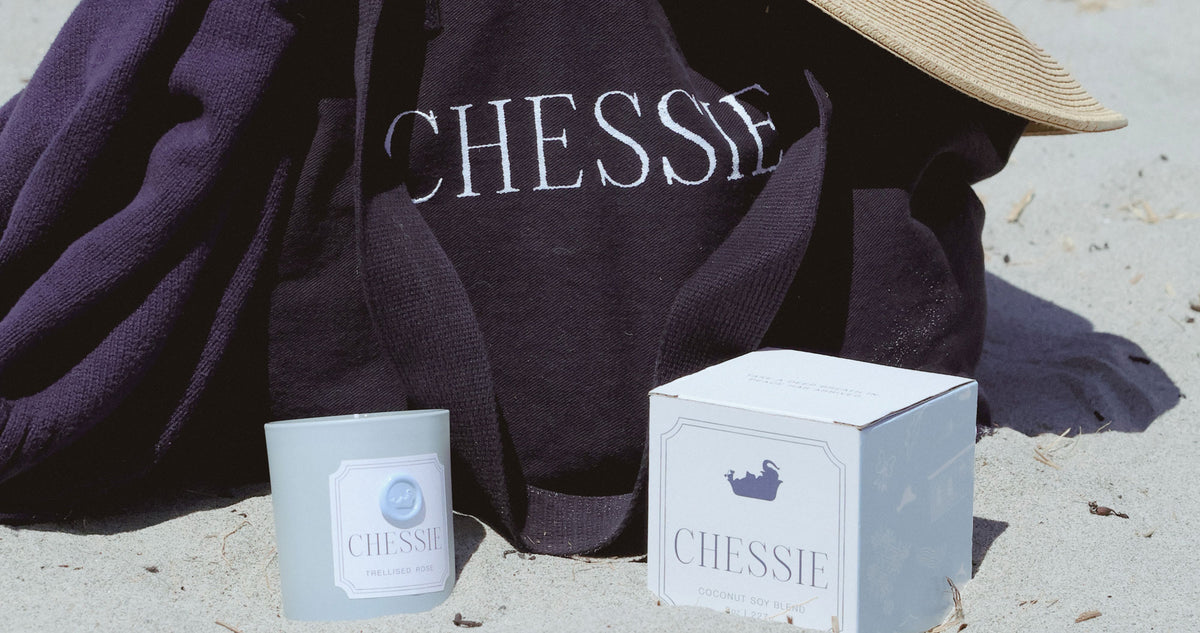 A dark navy beach tote with the word 'CHESSIE' printed in white, styled on a sandy beach next to a white product box and a small glass jar candle.