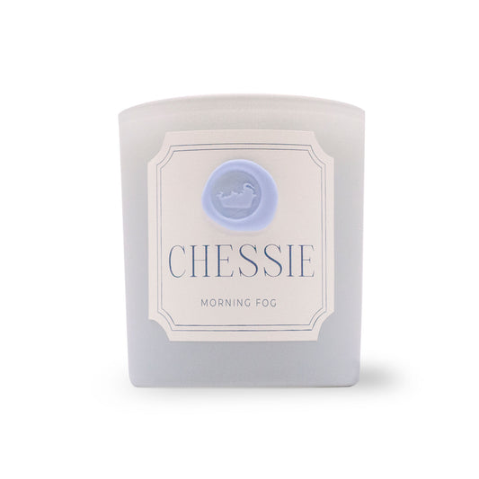 A frosted glass candle jar with a white label reading 'CHESSIE MORNING FOG' and a light blue circular logo, photographed on a white background.