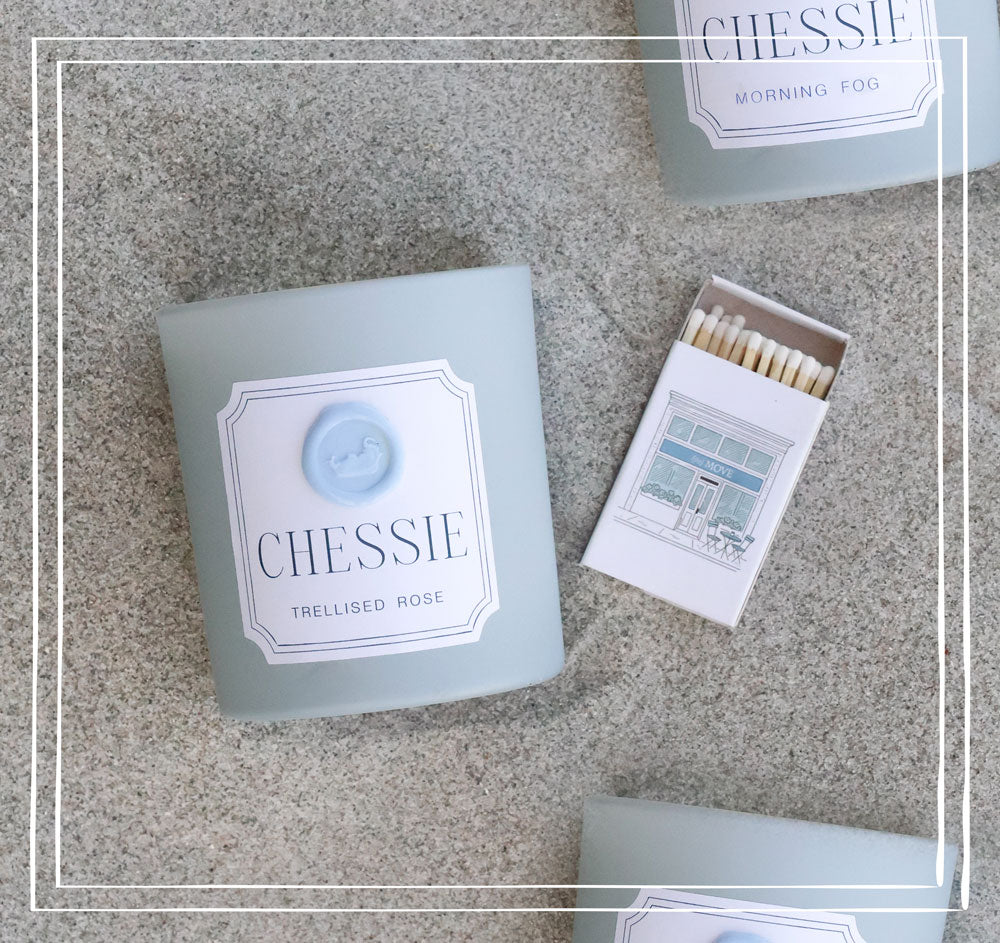 A styled flat-lay of CHESSIE candles and matchboxes on the beach. The main product shown is a square candle in a pale powder blue container with a white label reading 'CHESSIE TRELLISED ROSE' and a light blue wax seal.