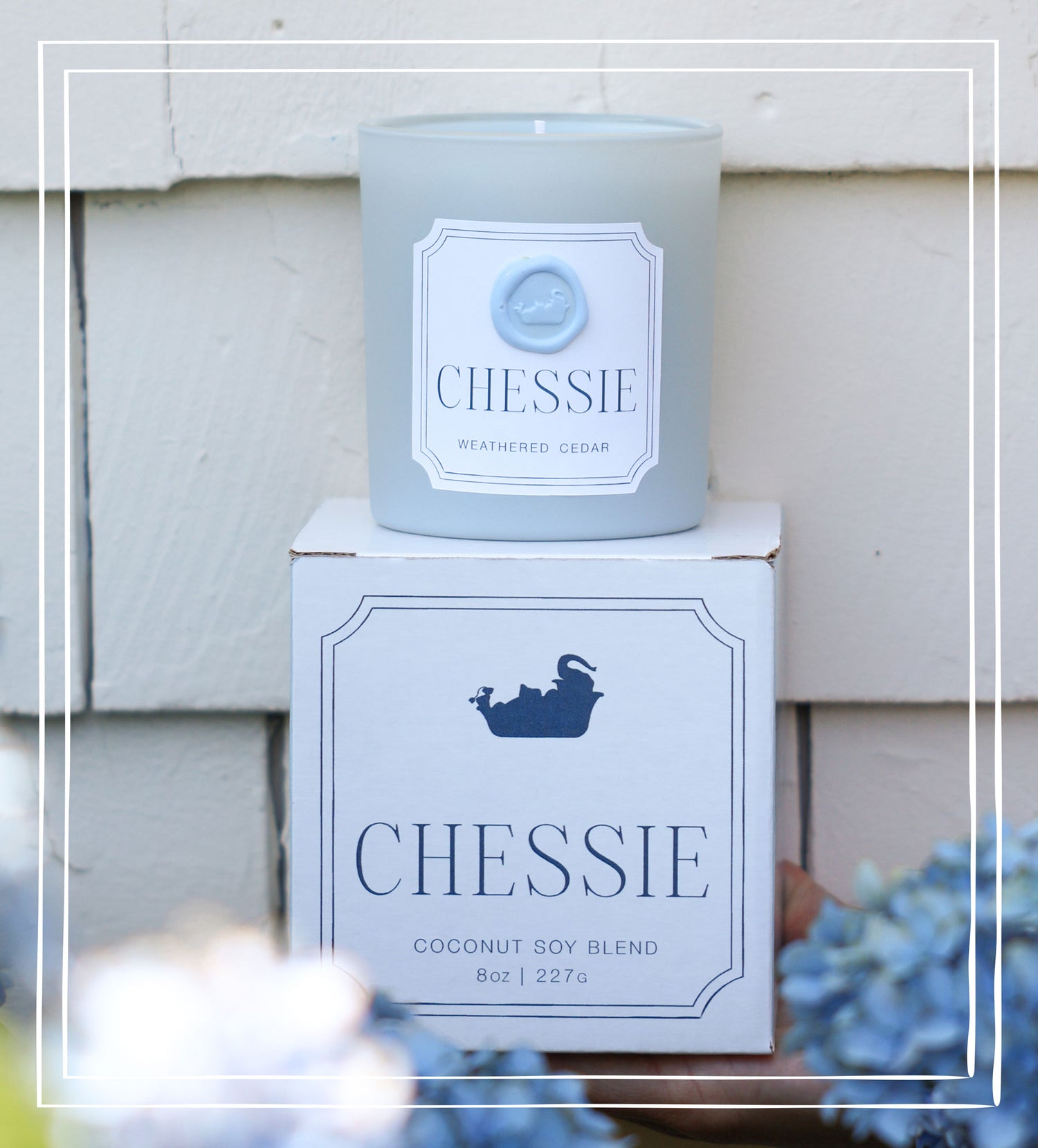A frosted light blue candle labeled 'CHESSIE' sits on top of its matching white and navy box in front of a white siding wall. The candle label reads 'WEATHERED CEDAR' and the box reads 'CHESSIE' with a small ship icon and 'COCONUT SOY BLEND 8oz | 227g'.