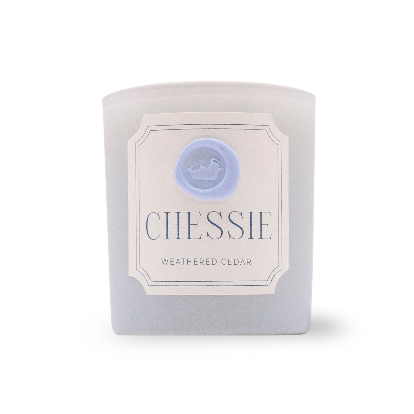 A frosted glass candle jar with a white label reading 'Chessie' and 'Weathered Cedar' and a light blue wax seal of a bathing elephant logo, photographed on a white background.